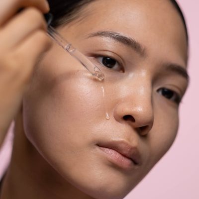 pexels-photo-8140913-8140913 Close-up of a woman applying skincare serum with a dropper against a pink background.